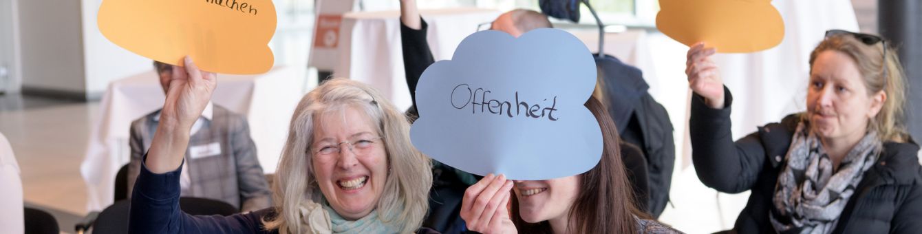 Menschen sitzen in Stuhlreihen und halten Wolken aus Papier hoch. Auf den Wolken der beiden Frauen in der ersten Reihe stehen "Offenheit" und "weiter machen"