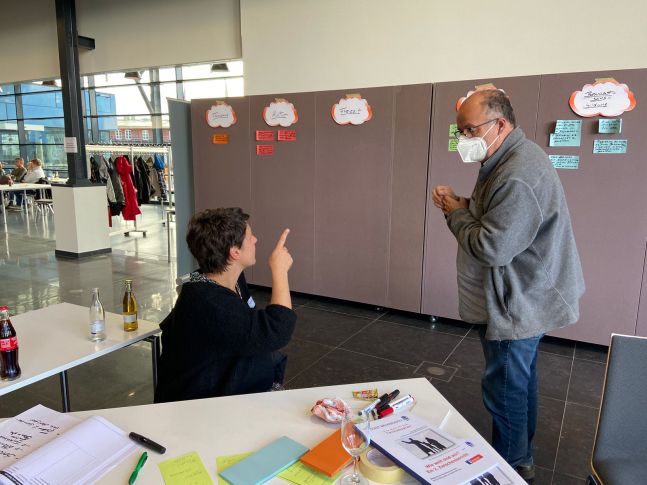 Ein Mann und eine Frau schauen auf die Arbeitsergebnisse einer Arbeitsgruppe, die auf Karten an eine Wand  geklebt wurden
